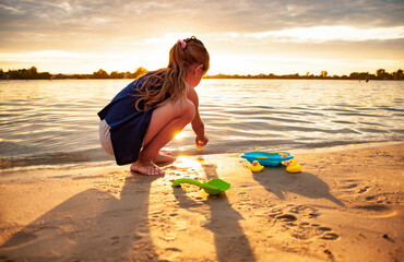 Side view of little lovely caucasian girl playing with tiny rubber yellow ducks in small blue pool, standing on beach sand. Attractive kid having fun, enjoying holidays at sea, summertime.