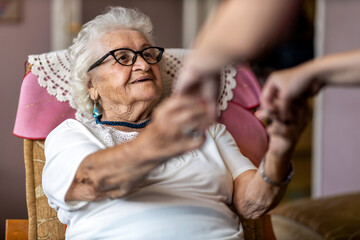 Female home carer supporting old woman to stand up from the armchair at care home