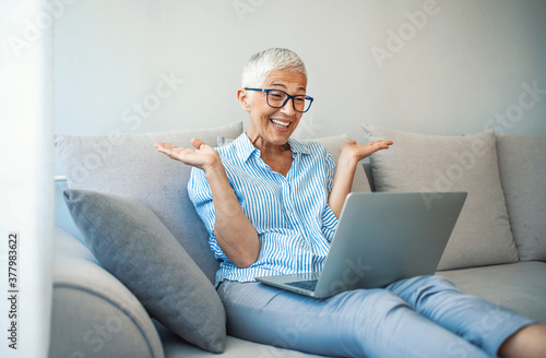 Technology, people and communication concept - Happy senior woman with tablet pc computer having video chat at home. Modern middle-aged 50s grandmother sit relax on couch in living room using laptop