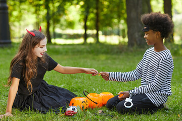 Wall Mural - Side view portrait of boy and girl wearing Halloween costumes and sharing candy while sitting on green grass outdoors