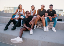 Group Of Young Modern African Black Friends Happily Sitting Together On The Bench Of The Stairs In Bright Sunlight Enjoying Summer Vacation