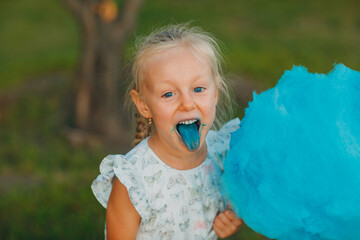 Wall Mural - Little blond girl eating cotton candy and shows blue tongue in the park.