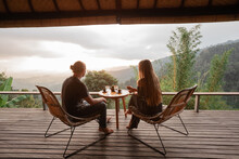 Happy Couple Sit On Wicker Chairs At Outdoor Terrace In The Mountains And Drink Wine. Romantic Time Together