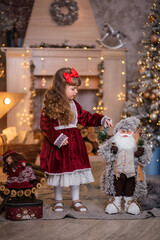 Wall Mural - Little beautiful girl in a red dress near the Christmas tree. Girl with a doll are playing Christmas. The child smiles. Beautiful girl with long curly hair. child in santa claus clothes