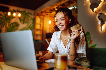 Young woman holding credit card and using laptop computer. African American woman sitting at cafe making online shopping. Online shopping, e-commerce, internet banking, spending money.