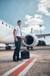 Good-looking male pilot with travel suitcase standing in airfield