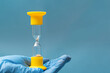 © Alena - A hand in a blue protective glove holds an hourglass on a blue background, close-up, copy space
