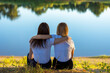 © NataliaSavilova - Two young women rear view hugging while sitting on the grass near the river in autumn. Friends leaned on each other in the park