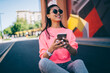© BullRun - Toothy hipster girl in stylish sunglasses enjoying leisure for networking smartphone browser, prosperous Asian millennial 20s with cellphone technology resting in city sitting at asphalt road