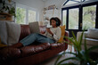 © StratfordProductions - African American young woman relaxing on couch listening to music while making online payment using bank card and laptop