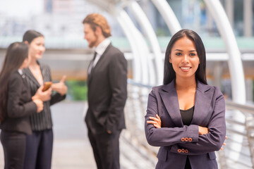 Beautiful young grinning professional Black woman folded arms and confident expression while standing outdoors with Business team standing together in the background.