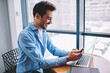 © BullRun - Successful male IT professional working remotely using smartphone gadget for sharing multimedia to laptop computer, happy Caucasian man smiling while reading received newsletter email message