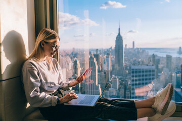 Wall Mural - Young distance worker sharing multimedia files via mobile and laptop technology connecting to bluetooth, Caucasian freelancer messaging on smartphone sitting at panoramic window in New York