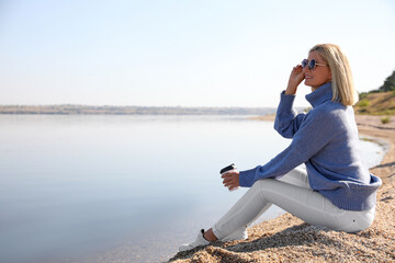 Poster - Happy woman in stylish sweater sitting on beach with cup of coffee