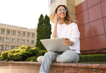 Wall Mural - Happy young woman with laptop talking on phone outdoors