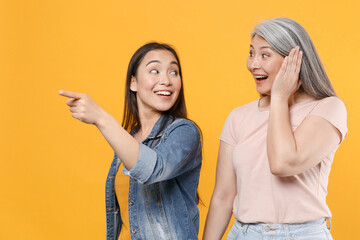 Canvas Print - Excited family asian women girls gray-haired mother and brunette daughter in casual clothes posing pointing index finger aside put hand on cheek isolated on yellow color background studio portrait.