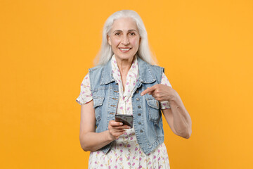 Smiling elderly gray-haired female woman wearing casual dress denim waistcoat posing pointing index finger on mobile cell phone typing sms message isolated on yellow color background studio portrait.