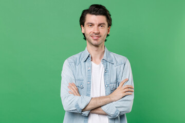 Smiling handsome attractive young brunet man 20s wearing casual clothes white t-shirt denim shirt posing holding hands crossed looking camera isolated on green color wall background studio portrait.