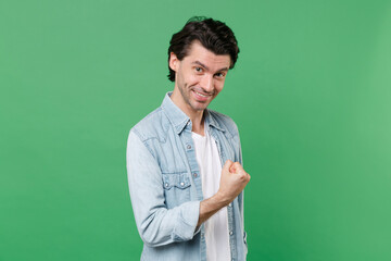 Smiling joyful young brunet man 20s wearing casual clothes white t-shirt denim shirt posing clenching fist doing winner gesture looking camera isolated on green color wall background studio portrait.