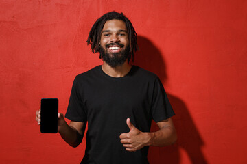 Wall Mural - Smiling young african american man 20s in black casual t-shirt posing hold mobile phone with blank empty screen mock up copy space showing thumb up isolated on red color background studio portrait.