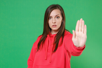 Wall Mural - Serious concerned young brunette woman 20s wearing bright red casual streetwear hoodie posing showing stop gesture with palm looking camera isolated on green color wall background studio portrait.