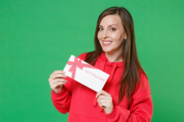 Wall Mural - Smiling beautiful young brunette woman 20s wearing bright red casual streetwear hoodie posing holding in hands gift certificate looking camera isolated on green color wall background studio portrait.