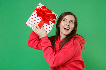 Wall Mural - Cheerful curious funny young brunette woman in red casual streetwear hoodie posing celebrating hold red present box with gift ribbon bow looking up isolated on green color background studio portrait.