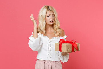 Wall Mural - Displeased young woman in white casual clothes hold red present box with gift ribbon bow spreading hands isolated on pink color background studio. Valentine's Day Women's Day birthday holiday concept.