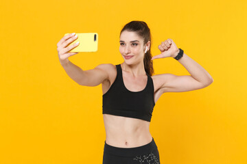 Wall Mural - Smiling young fitness sporty woman in black sportswear with air pods posing work out training doing selfie shot on mobile phone pointing thumb on herself isolated on yellow background studio portrait.