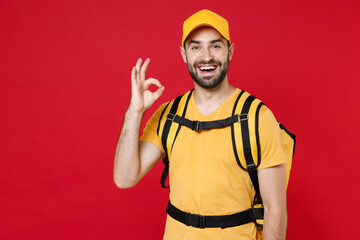 Wall Mural - Delivery employee man guy male 20s in yellow cap t-shirt uniform thermal bag backpack with food work as courier isolated on red background studio. Service during quarantine coronavirus covid-19 virus.