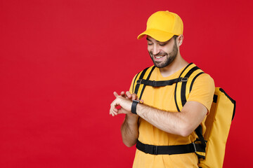 Wall Mural - Delivery man in yellow cap t-shirt uniform thermal bag backpack with food hand smartwatch isolated on red background studio Guy male employee work as courier Service coronavirus covid-19 virus concept