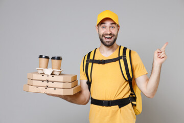 Wall Mural - Delivery employee man in yellow cap t-shirt uniform thermal food bag backpack work courier service on quarantine coronavirus covid-19 hold cardboard tea cups pizza flatbox isolated on gray background.