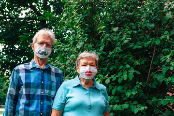 Elderly couple stands against a background of green foliage in disposable masks with mustaches and lips drawn by grandchildren. International Day of Older Persons and Grandparents during coronavirus