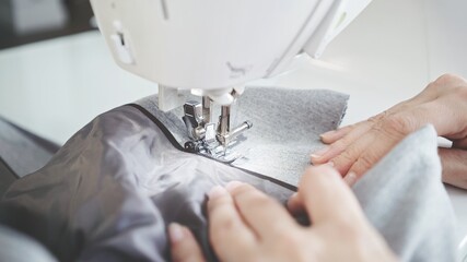 Concept of sewing in modern bright studio, woman in white sweater sewing grey cloth in process. Female hands sew on a white sewing machine close-up. 