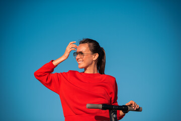 Cute smiling woman in red stylish top and sunglasses isolated on blue sky background holding handlebar of electric scooter and looking forward.