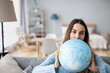 © BestForYou - Smiling young woman holding earth globe sitting on the couch at home. Planning vacation.
