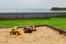Toys In A Children's Sandbox Fenced With A Wooden Fence On A Rainy Summer Day Against The Sea, Summer Holidays.
