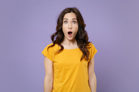 Shocked amazed surprised excited young brunette woman 20s wearing basic yellow t-shirt posing standing keeping mouth open looking camera isolated on pastel violet colour background, studio portrait.