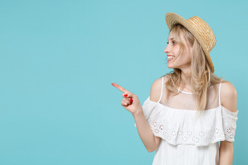 Wall Mural - Smiling attractive cute young blonde woman 20s wearing white summer dress hat standing pointing index finger aside on mock up copy space isolated on blue turquoise colour background studio portrait.