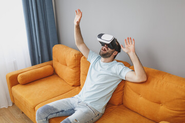 Wall Mural - Excited cheerful funny young bearded man wearing casual blue t-shirt watching in vr headset gadget spreading hands keeping mouth open sitting on couch resting spending time in living room at home.