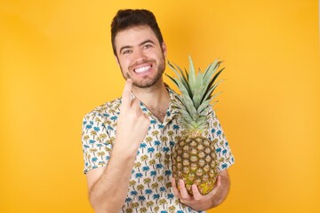 Young man holding pineapple wearing hawaiian shirt over yellow isolated background Beckoning come here gesture with hand inviting welcoming happy and smiling