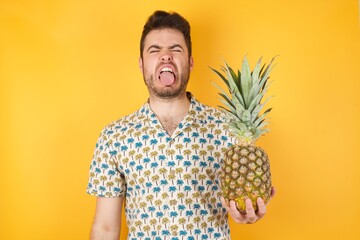 Young man holding pineapple wearing hawaiian shirt over yellow isolated background  sticking tongue out happy with funny expression. Emotion concept.