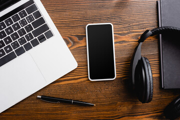 top view of smartphone with blank screen, laptop, wireless headphones, notebook and pen on office desk