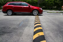 Red Minivan Driving Up To And Just Connecting With Yellow And Black Striped Speed Bump In Parking Lot With Diagonal Striped Spaces And Trees In The Background, Mid-Day