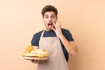 Male baker holding a table with several breads isolated on beige background with surprise facial expression