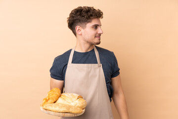 Wall Mural - Male baker holding a table with several breads isolated on beige background looking to the side