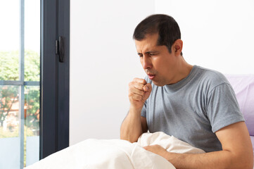 Shot of a handsome young man suffering with thecoughing as symptom for cold or bronchitis while sitting on his bed at home