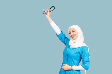 Wall Mural - a friendly, muslim doctor or nurse woman in hijab with a stethoscope on a blue background. emotions of victory or success