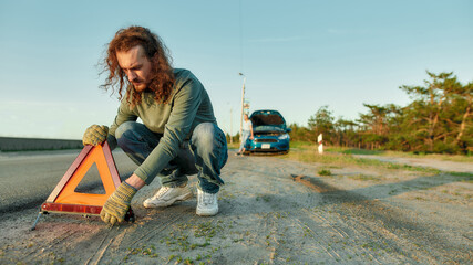 Wall Mural - Man driver putting red warning triangle or emergency stop sign behind his broken car on the side of the road, his girlfriend waiting in the background