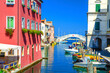 Stone bridge Ponte di Vigo across Vena water canal with colorful boats and old buildings in historical centre of Chioggia town, blue sky background in summer day, Veneto Region, Northern Italy